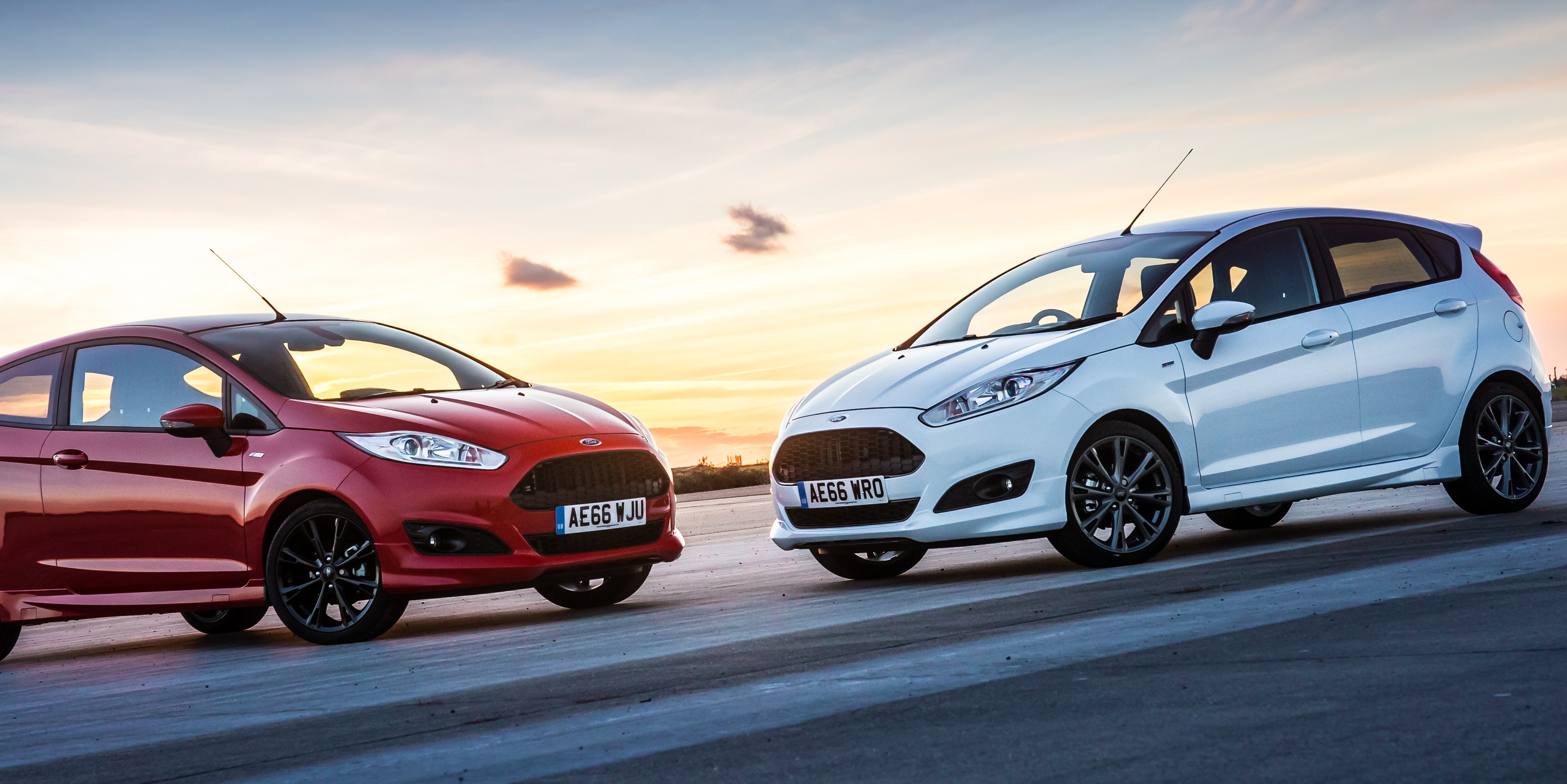 Red Ford Fiesta on left and white Ford Fiesta on right, nose to nose, with sunset in background